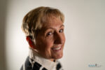 Smiling older woman with short blonde hair looks over her shoulder, wearing a striped shirt, against a plain background—her confident grin is a perfect example of how long do porcelain veneers last when well cared for.