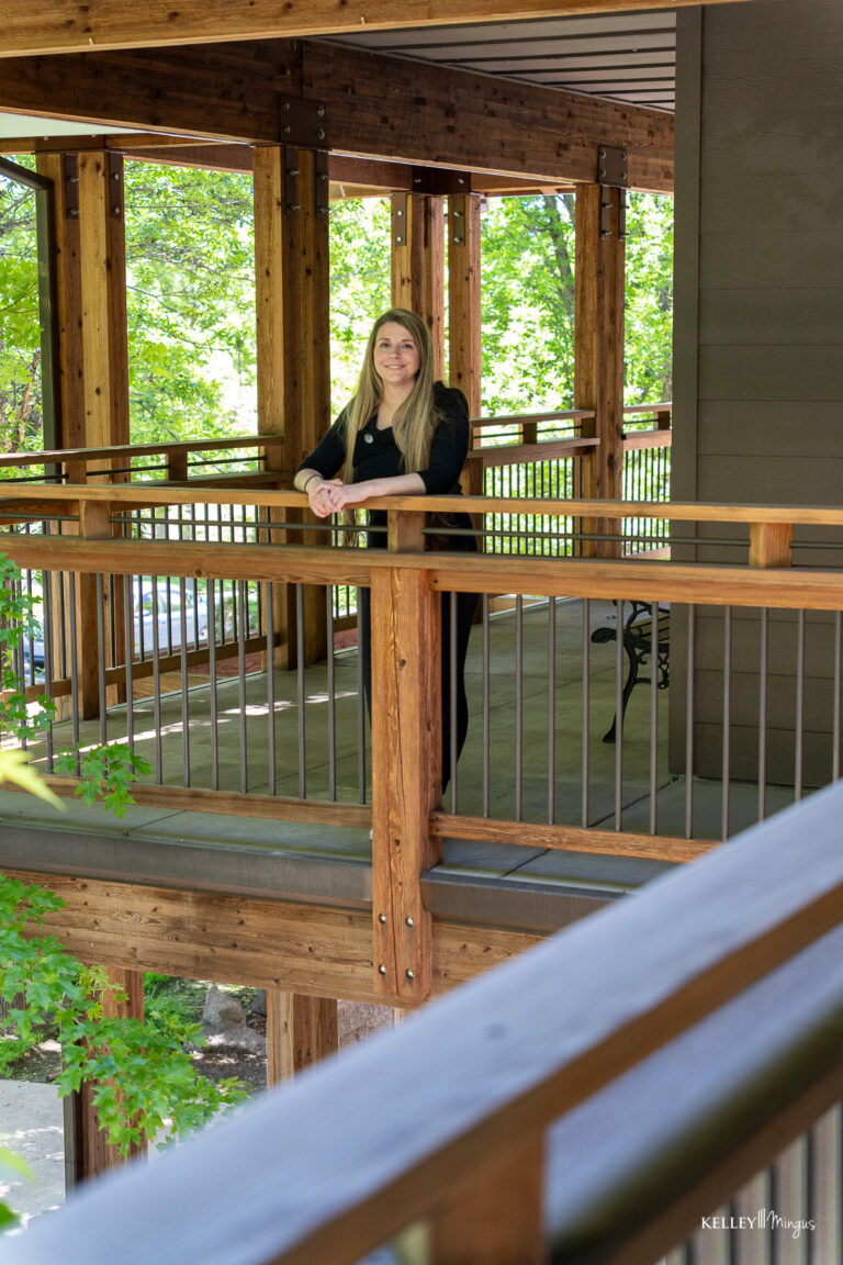 A woman with long blonde hair, wearing a black top and pants, leans on a wooden railing of an outdoor walkway surrounded by trees—enjoying the peaceful scenery near her trusted family dentist in Bend, Oregon.