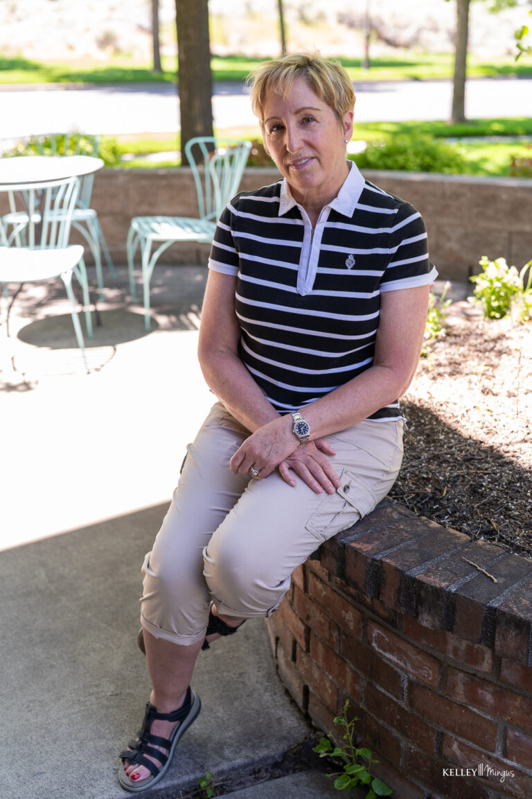 A person with short blond hair, wearing a black and white striped polo and beige pants, sits on a low brick wall outdoors near empty patio chairs—much like Kelley Mingus, sleep apnea specialist, unwinding after a long day.