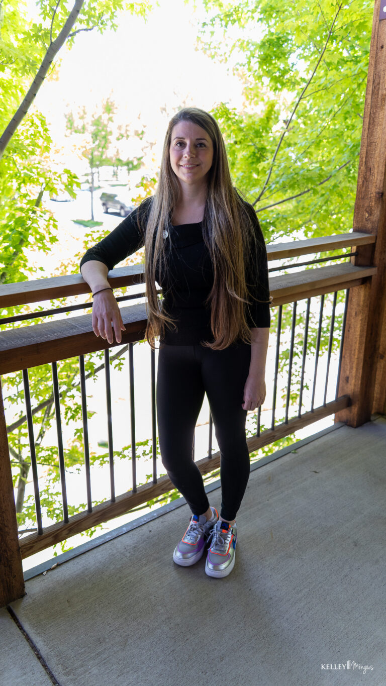 A person with long hair, dressed in black clothing and colorful sneakers, stands on a wooden balcony with green leaves and trees in the background, radiating a calm presence reminiscent of someone who appreciates holistic dentistry and natural wellness.