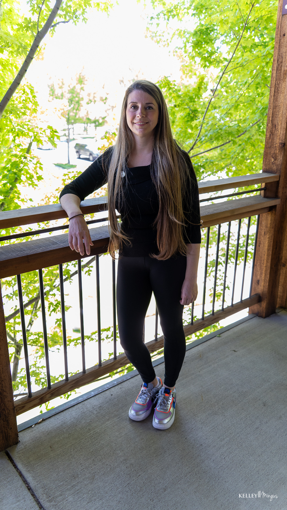 A person with long hair, dressed in black clothing and colorful sneakers, stands on a wooden balcony with green leaves and trees in the background, radiating a calm presence reminiscent of someone who appreciates holistic dentistry and natural wellness.