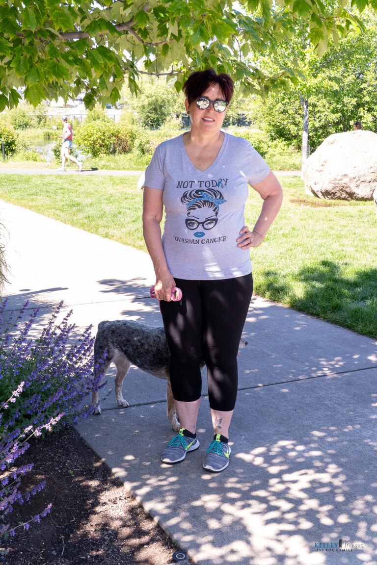 Woman stands on a sidewalk under a tree, wearing sunglasses and a “Not Today Ovarian Cancer” T-shirt, holding a pink item, with a dog partially visible behind her—perhaps on her way to an appointment with her trusted Bend Oregon cosmetic dentist.