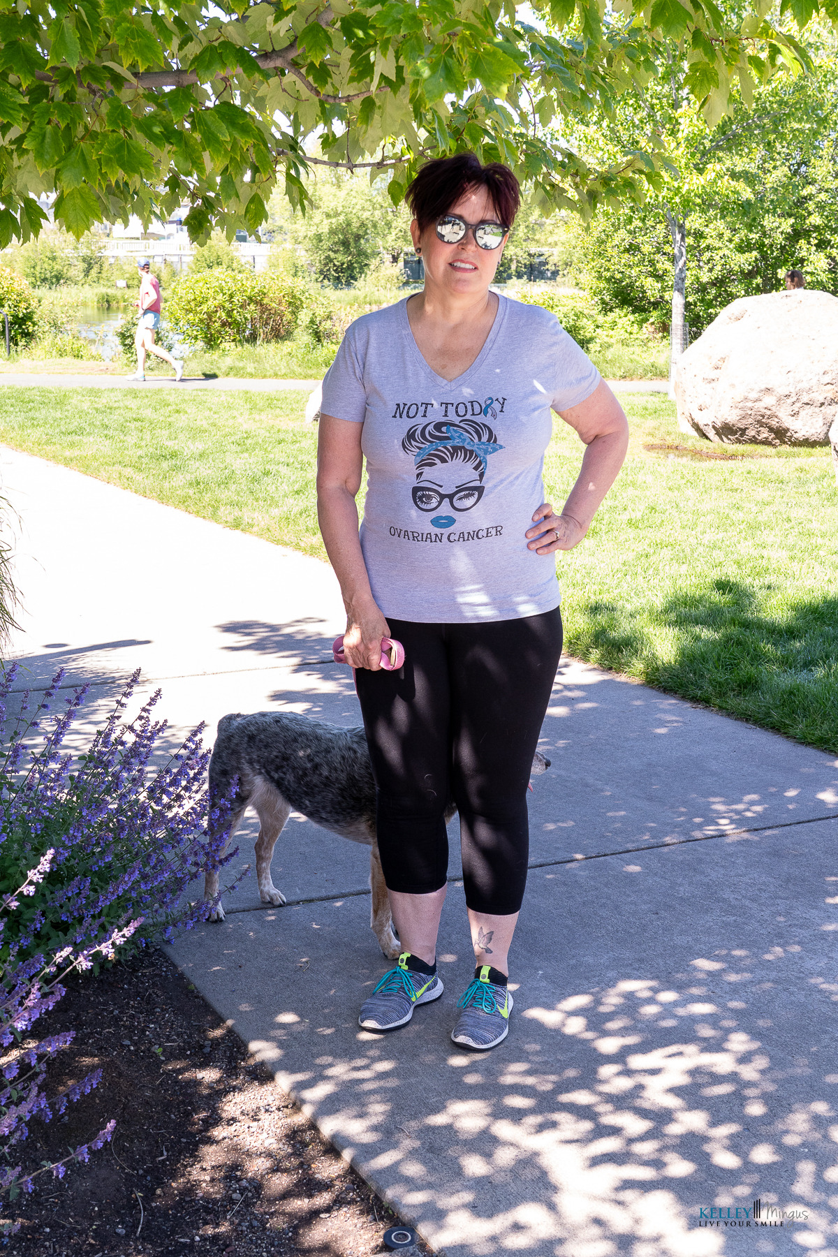 Woman stands on a sidewalk under a tree, wearing sunglasses and a “Not Today Ovarian Cancer” T-shirt, holding a pink item, with a dog partially visible behind her—perhaps on her way to an appointment with her trusted Bend Oregon cosmetic dentist.