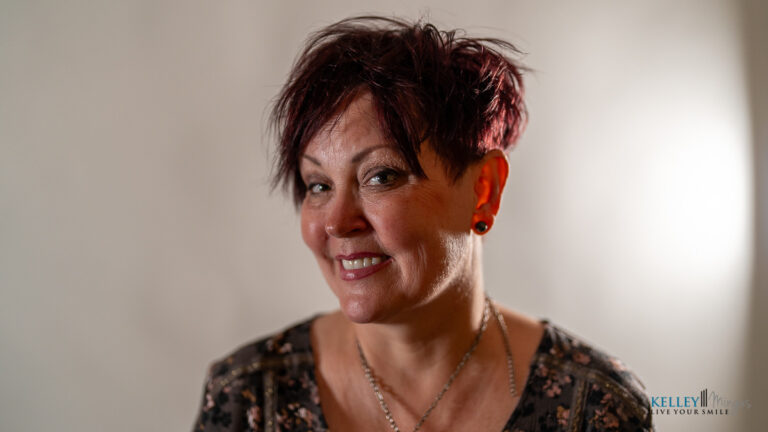 Woman with short, dark reddish hair and earrings smiling at the camera, wearing a patterned top, with a blurred neutral background—an approachable presence reminiscent of Kelley Mingus, sleep apnea specialist.