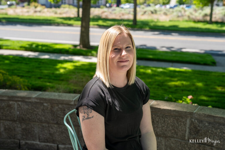 A woman with straight blonde hair and a black shirt sits outdoors on a metal chair, her upper left arm tattoo visible. A sidewalk and greenery, including a nearby holistic dentistry office, form the background.