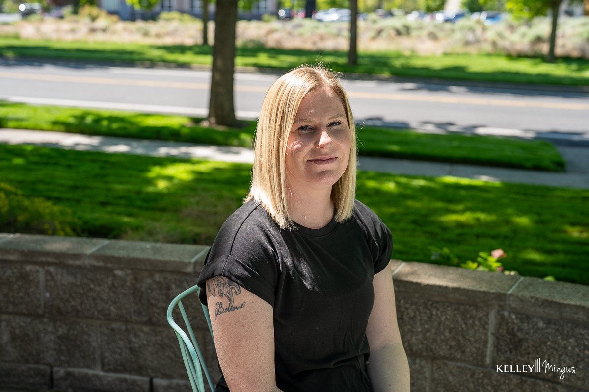 A woman with straight blonde hair and a black shirt sits outdoors on a metal chair, her upper left arm tattoo visible. A sidewalk and greenery, including a nearby holistic dentistry office, form the background.