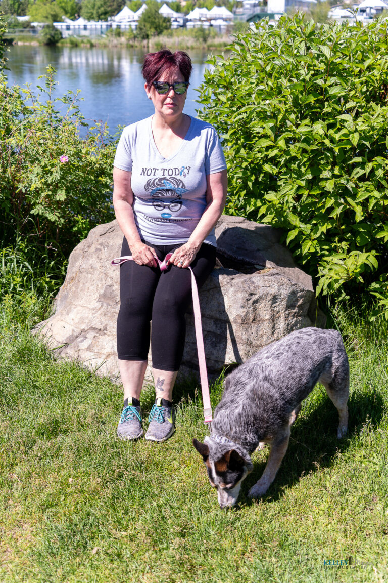 A woman sits on a rock by a lake holding a leash, while a gray dog sniffs the grass nearby on a sunny day, reflecting on the differences between full mouth rehabilitation vs. smile makeover as she enjoys the peaceful scenery.