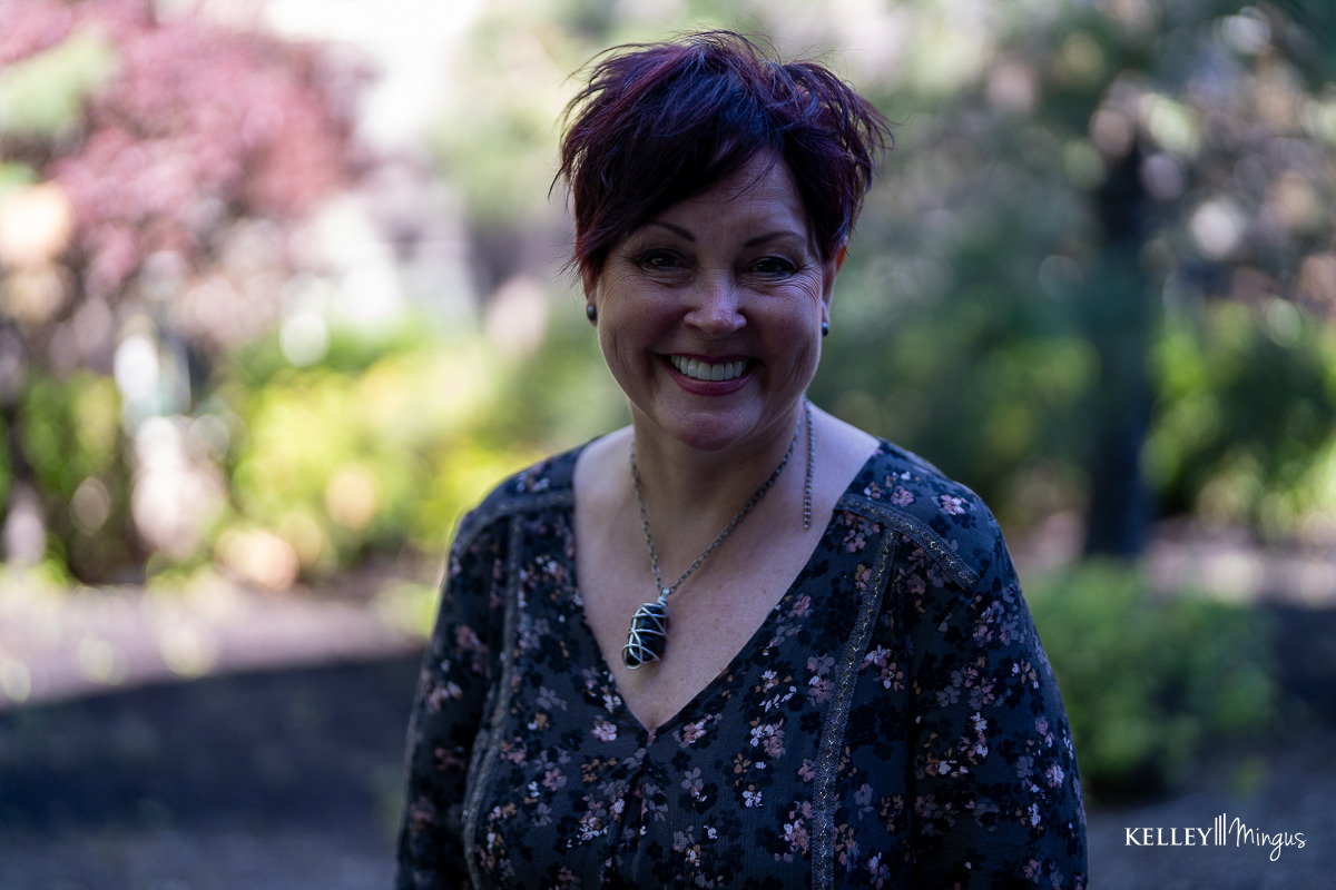 A woman with short dark hair and a floral blouse smiles outdoors in a garden setting, radiating the confidence of someone who’s just visited a Bend Oregon cosmetic dentist.