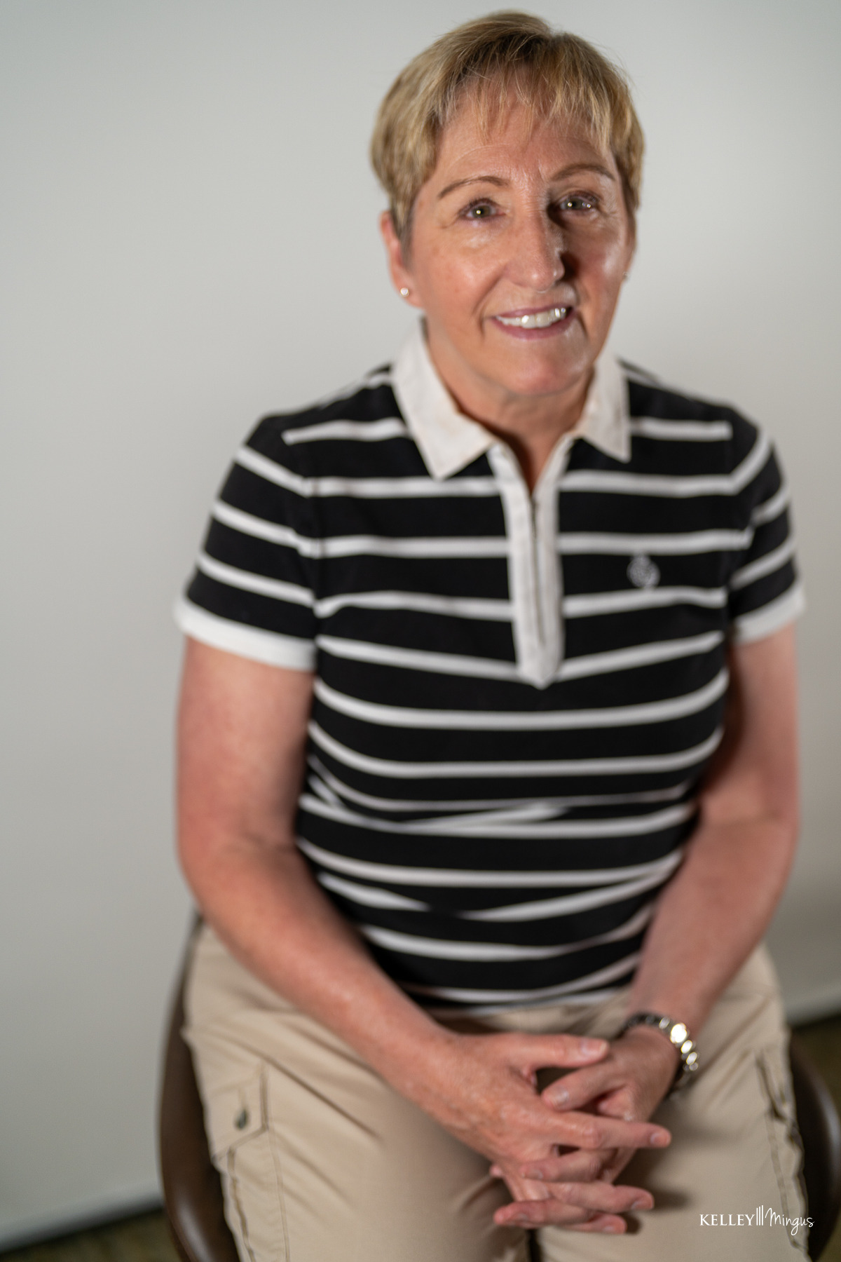 A person with short light hair, wearing a black-and-white striped polo shirt and khaki pants, sits on a stool against a plain background with hands folded in their lap, as if ready to discuss topics like full mouth rehabilitation vs. smile makeover.