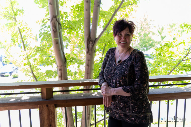 A woman with short dark hair stands and smiles on a wooden balcony, with green trees and sunlight in the background, reminiscent of content patients helped by Kelley Mingus, sleep apnea specialist.