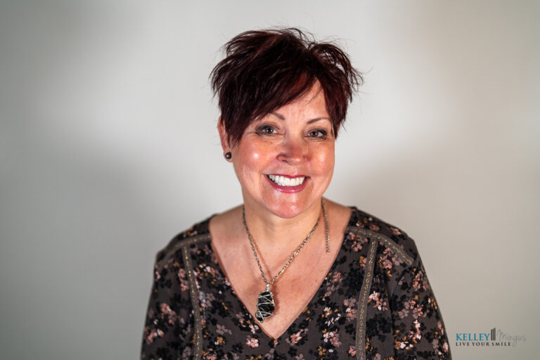 A woman with short, dark hair and a floral blouse smiles at the camera against a plain, light background. She is wearing a necklace and earrings, showcasing a radiant smile that subtly hints at the confidence one might gain from knowing how long porcelain veneers last.