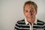 Smiling older woman with short blonde hair wearing a black and white striped collared shirt, standing in front of a plain light background—her genuine smile reminiscent of the care provided by Kelley Mingus, sleep apnea specialist.