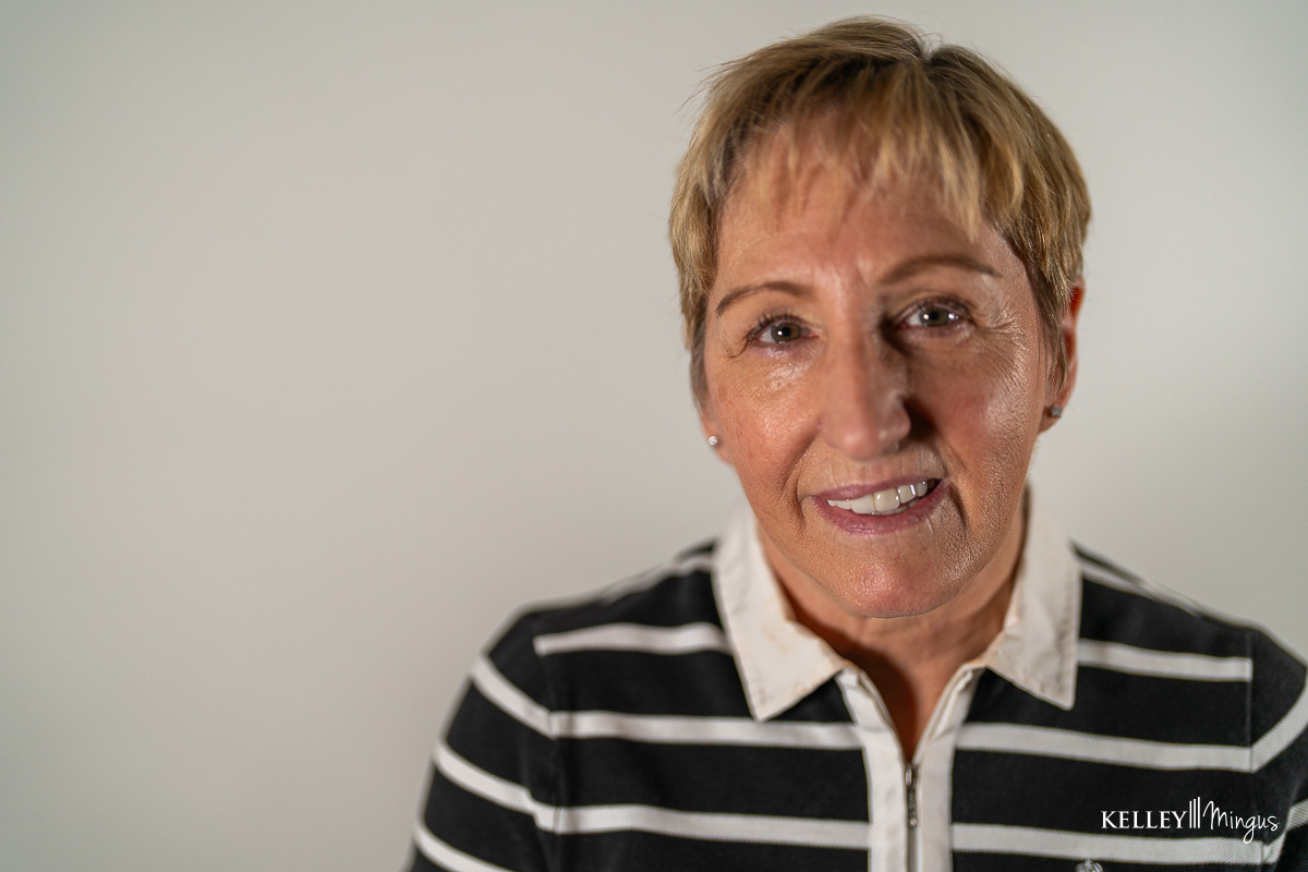 Smiling older woman with short blonde hair wearing a black and white striped collared shirt, standing in front of a plain light background—her genuine smile reminiscent of the care provided by Kelley Mingus, sleep apnea specialist.