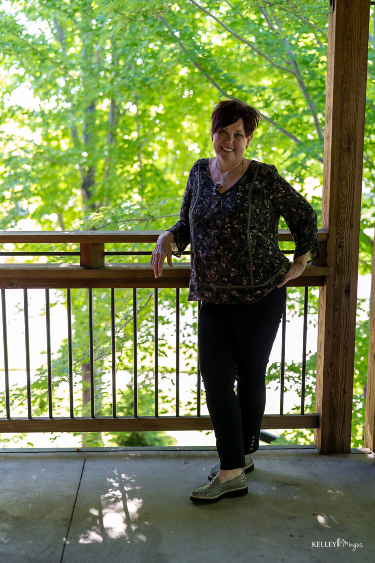 A woman stands on a shaded wooden deck, leaning against the railing, with green trees and sunlight in the background—her relaxed confidence reminiscent of someone who has discovered the difference between full mouth rehabilitation vs. smile makeover.