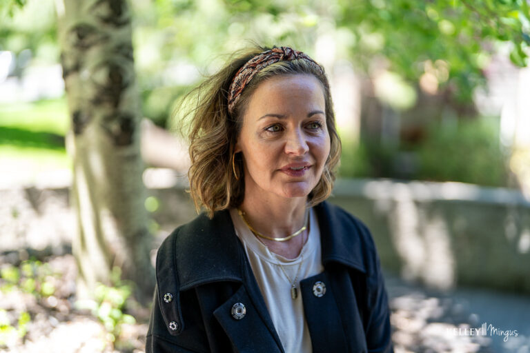 A woman with shoulder-length hair and a headband is sitting outdoors in a shaded area, wearing a dark jacket and layered necklaces, perhaps waiting for her appointment with her trusted family dentist in Bend, Oregon.