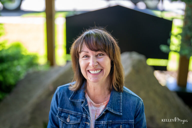 Woman with brown hair and bangs smiles at the camera, wearing a denim jacket and light top, outdoors with greenery and sunlight in the background, exuding confidence similar to someone who just learned how long do porcelain veneers last.
