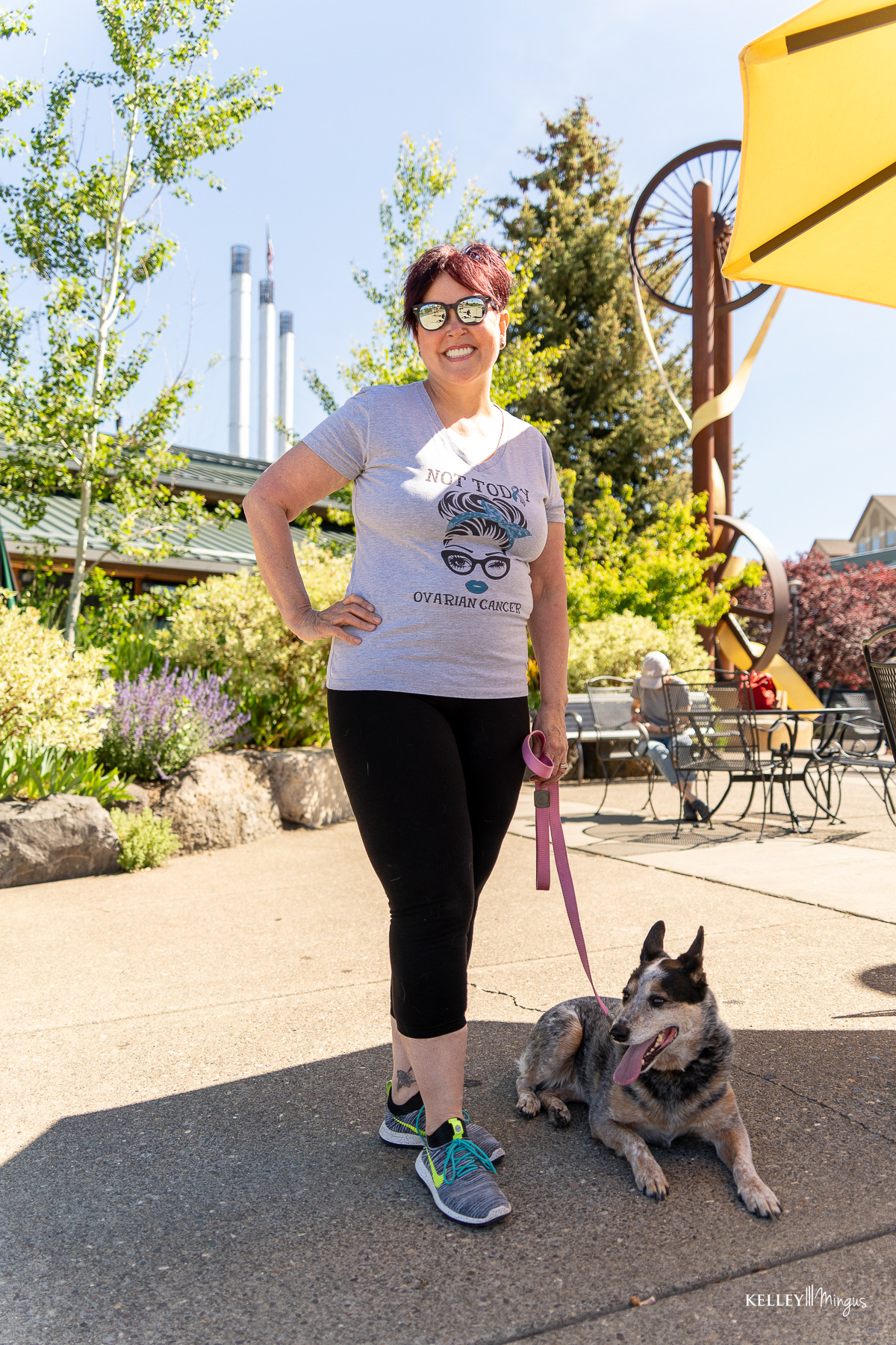 Woman in sunglasses stands on a sunny sidewalk holding a pink leash attached to a dog lying at her feet; outdoor seating and plants are visible in the background—a serene scene reminiscent of an afternoon outside Kelley Mingus sleep apnea specialist's office.