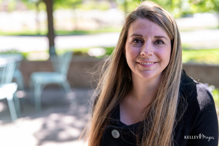 A woman with long blonde hair and a black top smiles at the camera while sitting outdoors near light blue chairs, enjoying a sunny day after her visit to a family dentist in Bend, Oregon.