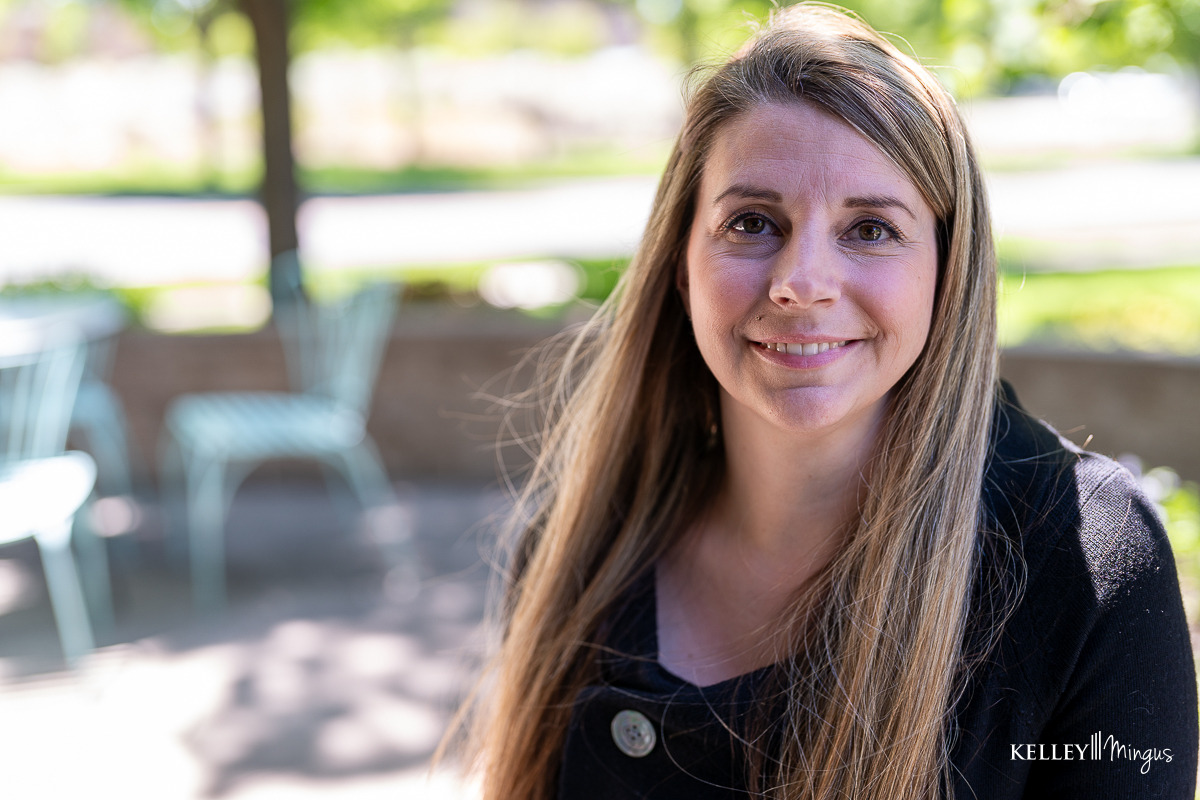 A woman with long blonde hair and a black top smiles at the camera while sitting outdoors near light blue chairs, enjoying a sunny day after her visit to a family dentist in Bend, Oregon.