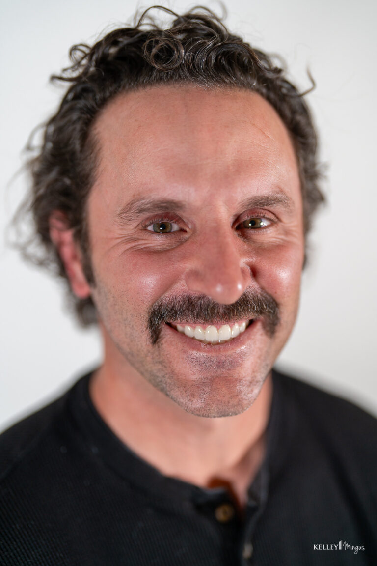 Man with short curly hair and a mustache smiling at the camera, wearing a black shirt, photographed against a plain white background—showcasing the confidence that comes with affordable cosmetic dentistry in Bend.