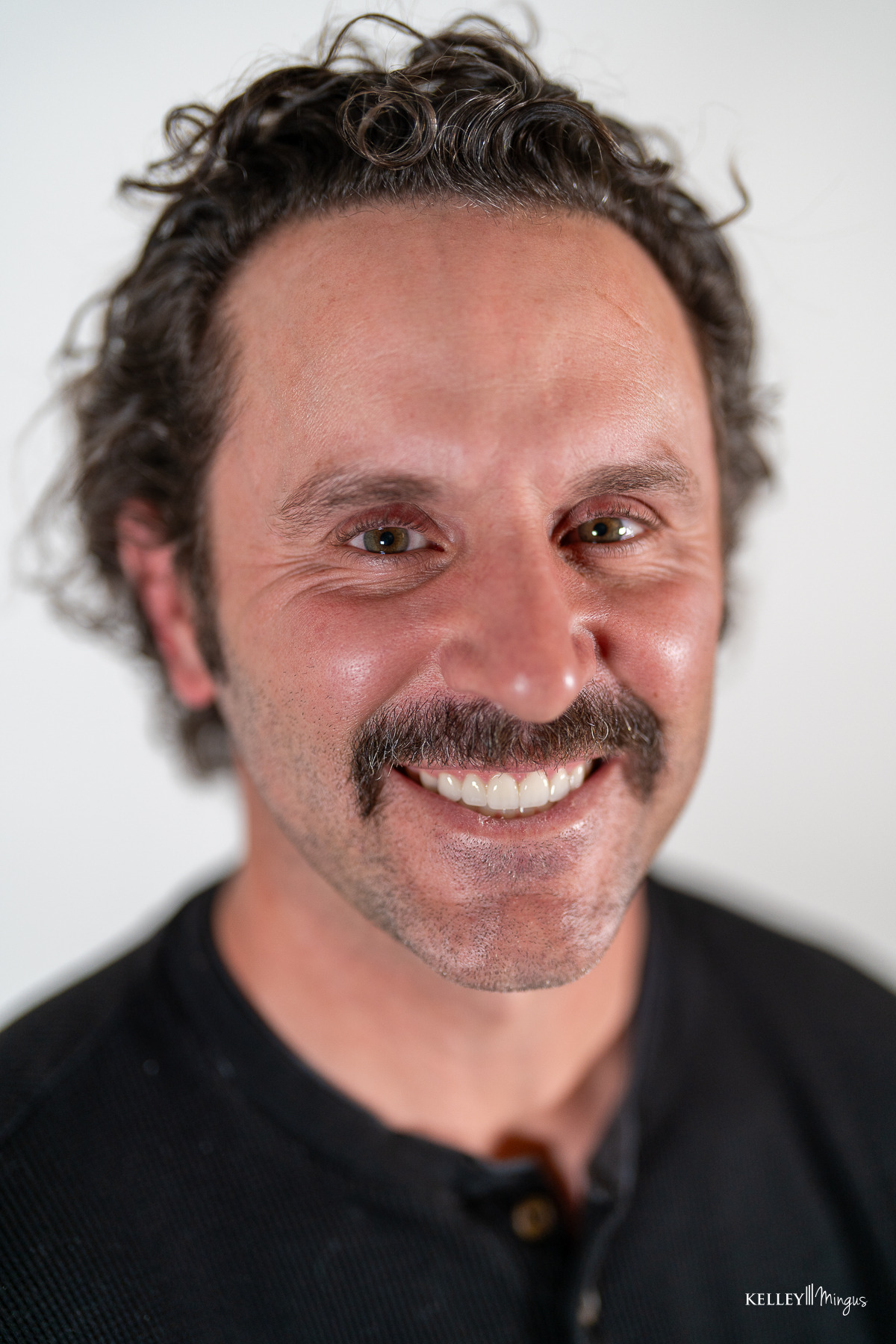 Man with short curly hair and a mustache smiling at the camera, wearing a black shirt, photographed against a plain white background—showcasing the confidence that comes with affordable cosmetic dentistry in Bend.