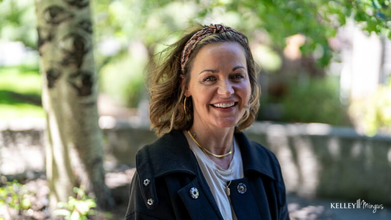 Woman with shoulder-length hair and a headband smiles outdoors, wearing a dark jacket and layered necklaces—perhaps enjoying a break after visiting her trusted family dentist in Bend, Oregon. Sunlight filters through green foliage in the background.
