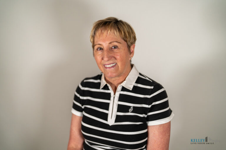 A woman with short, light brown hair wears a black and white striped shirt and smiles at the camera in front of a plain background, her confident smile suggesting she knows just how long do porcelain veneers last.