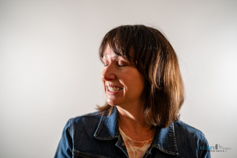 A woman with shoulder-length brown hair smiles with her eyes closed, showcasing the confidence that comes from affordable cosmetic dentistry in Bend, while wearing a denim jacket against a plain white background.