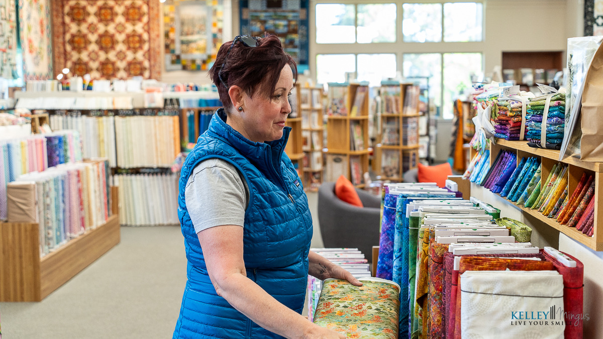 A woman stands in a fabric store, her smile bright from recent teeth cleaning and cosmetic dentistry, as she selects from colorful bolts of fabric displayed on shelves and tables around her.