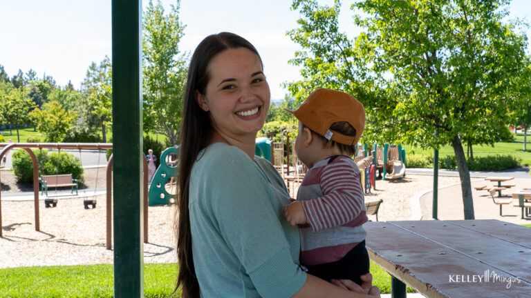 A woman smiles at the camera while holding a small child at an outdoor playground with green trees and play structures in the background, enjoying a carefree moment after visiting Kelley Mingus, TMJ specialist.