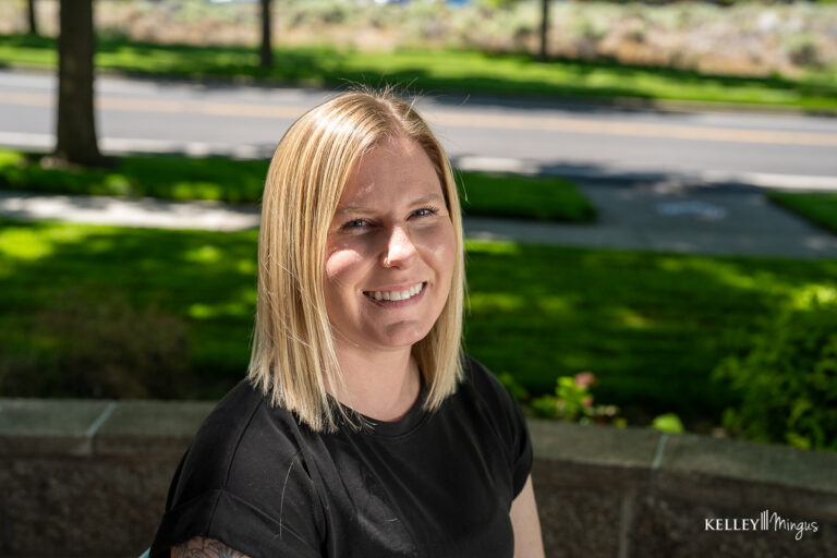 A person with straight blond hair and a black shirt smiles outdoors, sitting on a stone bench with a street and greenery in the background, radiating the confidence that often accompanies holistic dentistry.