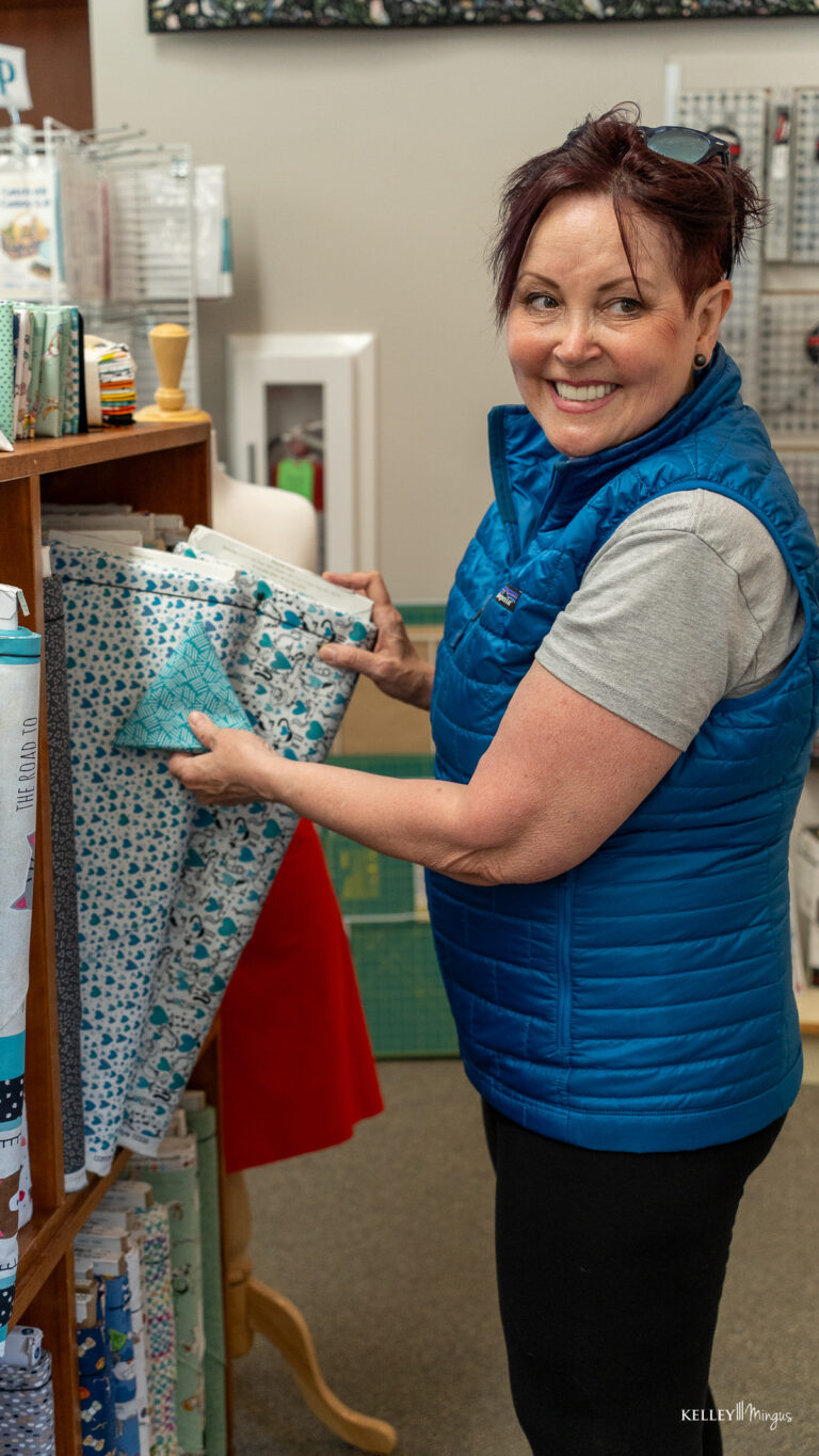 A woman wearing a blue vest smiles while selecting patterned fabric from a shelf in a fabric store, her bright smile reminiscent of a Bend, Oregon cosmetic dentist’s expert touch.
