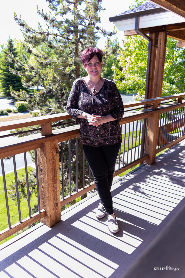 A person with short red hair stands on a wooden balcony, leaning on the railing, with trees and sunlight in the background, perhaps pondering everyday questions like how long do porcelain veneers last.