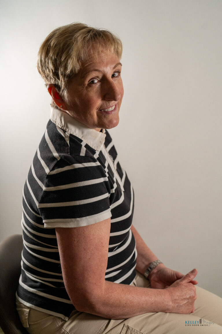 A person with short blonde hair, wearing a black and white striped shirt, sits sideways on a chair against a plain light background, looking toward the camera—composed and attentive, much like someone awaiting advice from Kelley Mingus, a renowned sleep apnea specialist.