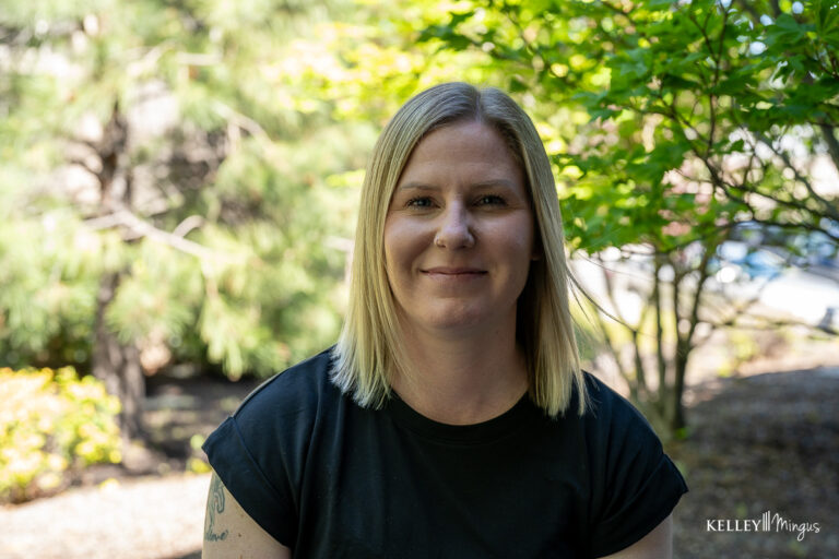 A woman with straight blonde hair and a black t-shirt sits outdoors in front of green foliage, smiling slightly at the camera—her radiant smile reflecting the confidence that comes from holistic dentistry.