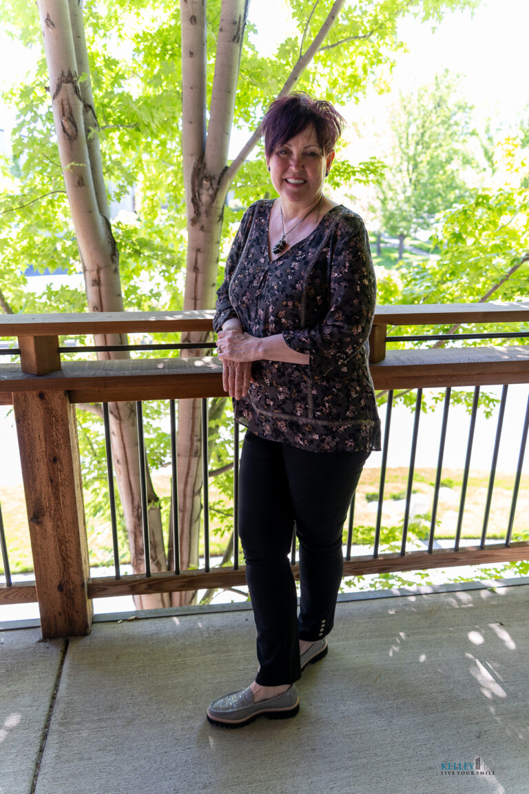 A woman with short dark hair stands on a balcony, smiling warmly. She wears a patterned blouse, black pants, and slip-on shoes—appearing both comfortable and confident, much like Kelley Mingus TMJ specialist. Trees and sunlight are visible in the background.