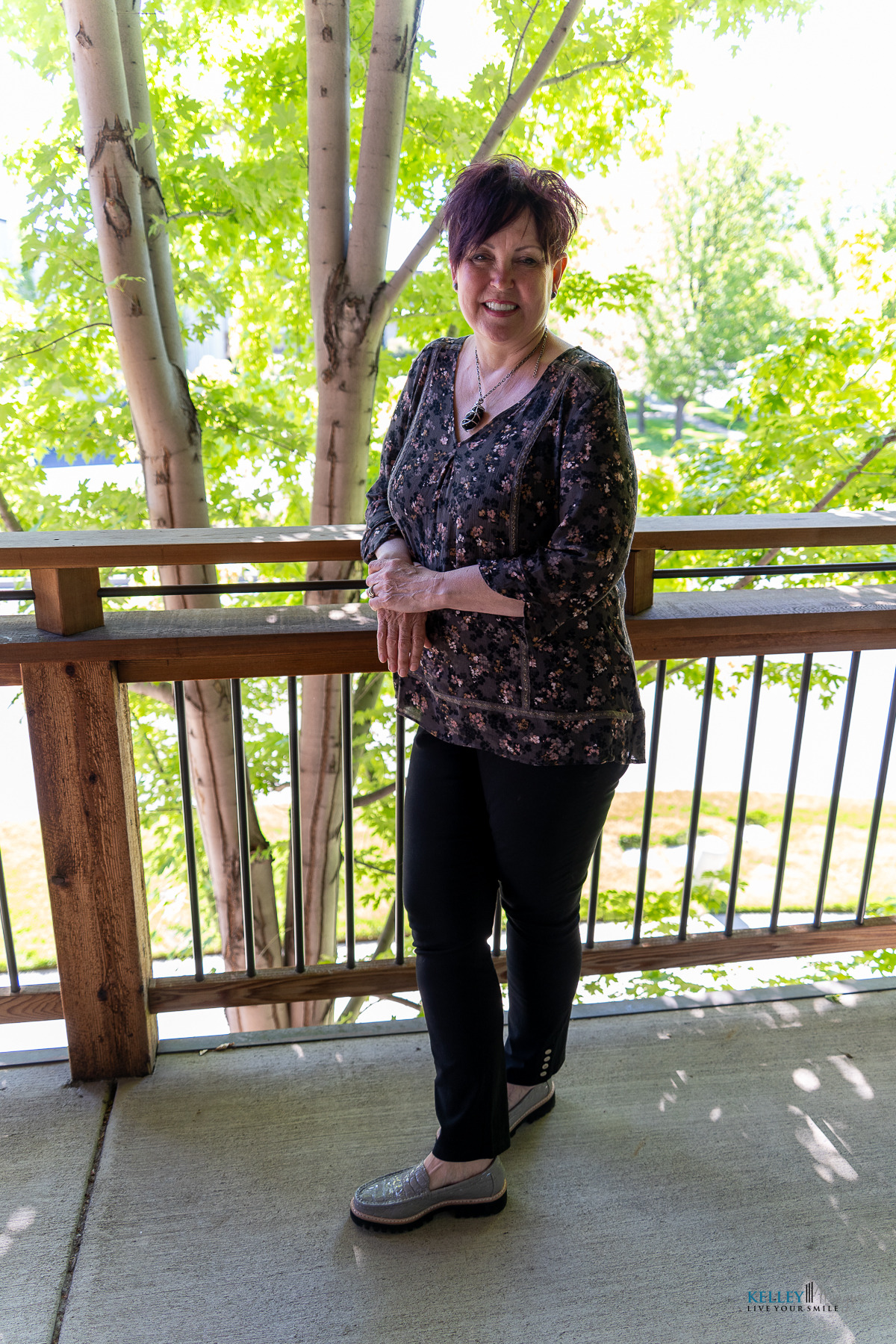 A woman with short dark hair stands on a balcony, smiling warmly. She wears a patterned blouse, black pants, and slip-on shoes—appearing both comfortable and confident, much like Kelley Mingus TMJ specialist. Trees and sunlight are visible in the background.