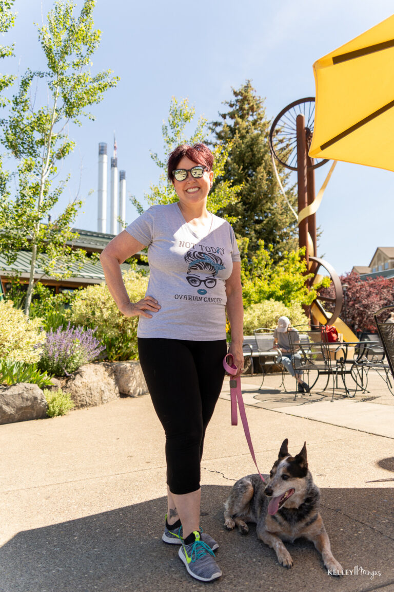 Woman in sunglasses stands outdoors holding a dog on a leash, with trees, yellow umbrellas, and a metal sculpture in the background on a sunny day—enjoying life’s simple moments after her Full Mouth Rehabilitation in Bend Oregon.