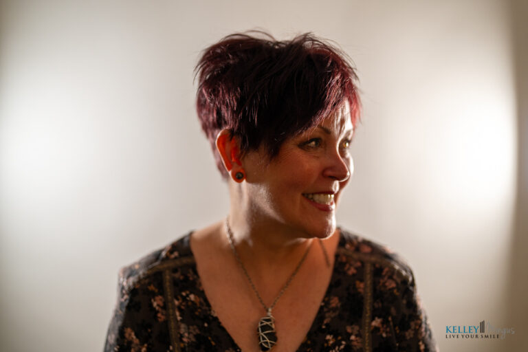 A woman with short, dark red hair smiles and looks to the side. She wears a patterned top, a pendant necklace, and red earrings—her bright smile reflecting the confidence of someone who recently visited a Bend Oregon cosmetic dentist. The background is softly lit and neutral.