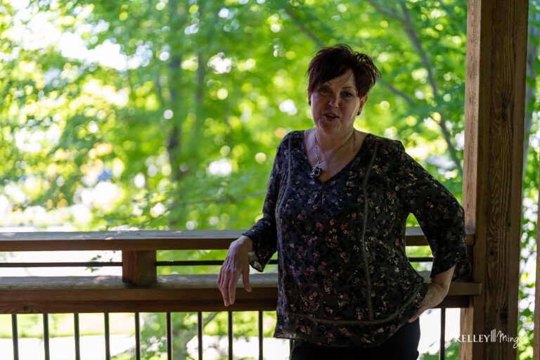A woman with short dark hair stands on a wooden porch, leaning against the railing, with green trees in the background—radiating confidence much like someone who has just completed Full Mouth Rehabilitation in Bend, Oregon.