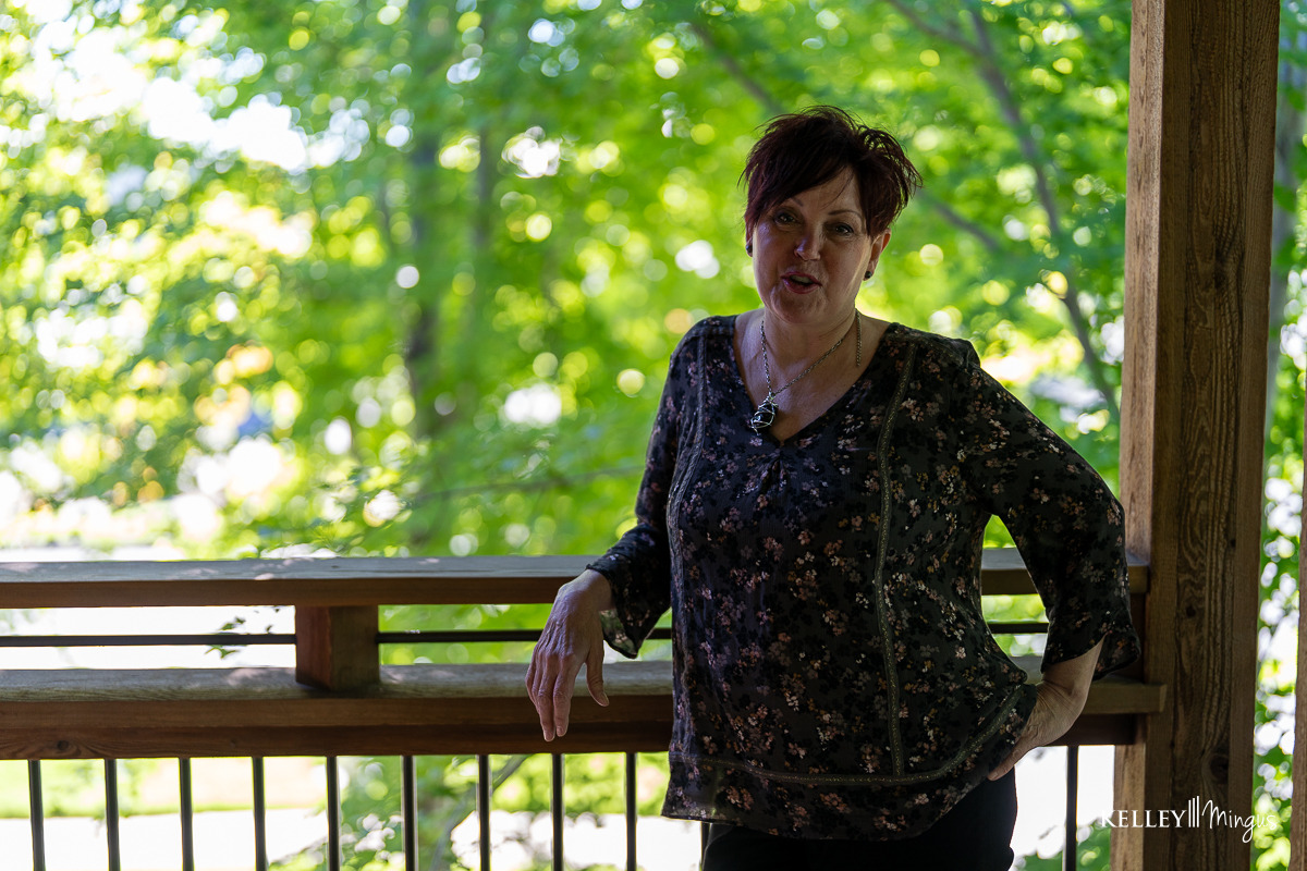 A woman with short dark hair stands on a wooden porch, leaning against the railing, with green trees in the background—radiating confidence much like someone who has just completed Full Mouth Rehabilitation in Bend, Oregon.