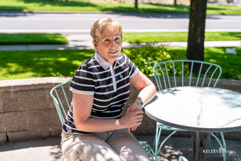 A woman with short hair, who recently visited a clinic for teeth cleaning and cosmetic dentistry, sits at an outdoor table on a sunny day, wearing a black and white striped shirt and beige pants.