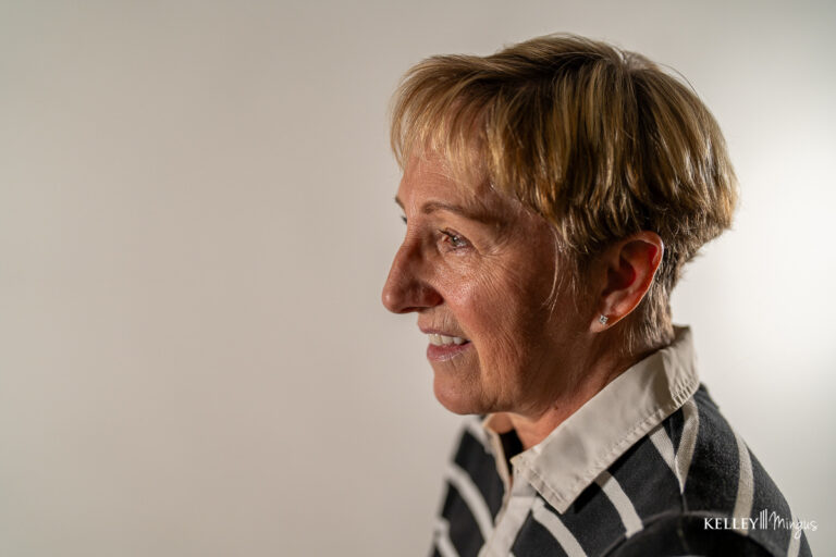 An older woman with short blond hair smiles, looking to the side, her bright smile reflecting confidence after teeth cleaning and cosmetic dentistry. She wears a striped shirt with a white collar. The background is plain and light-colored.
