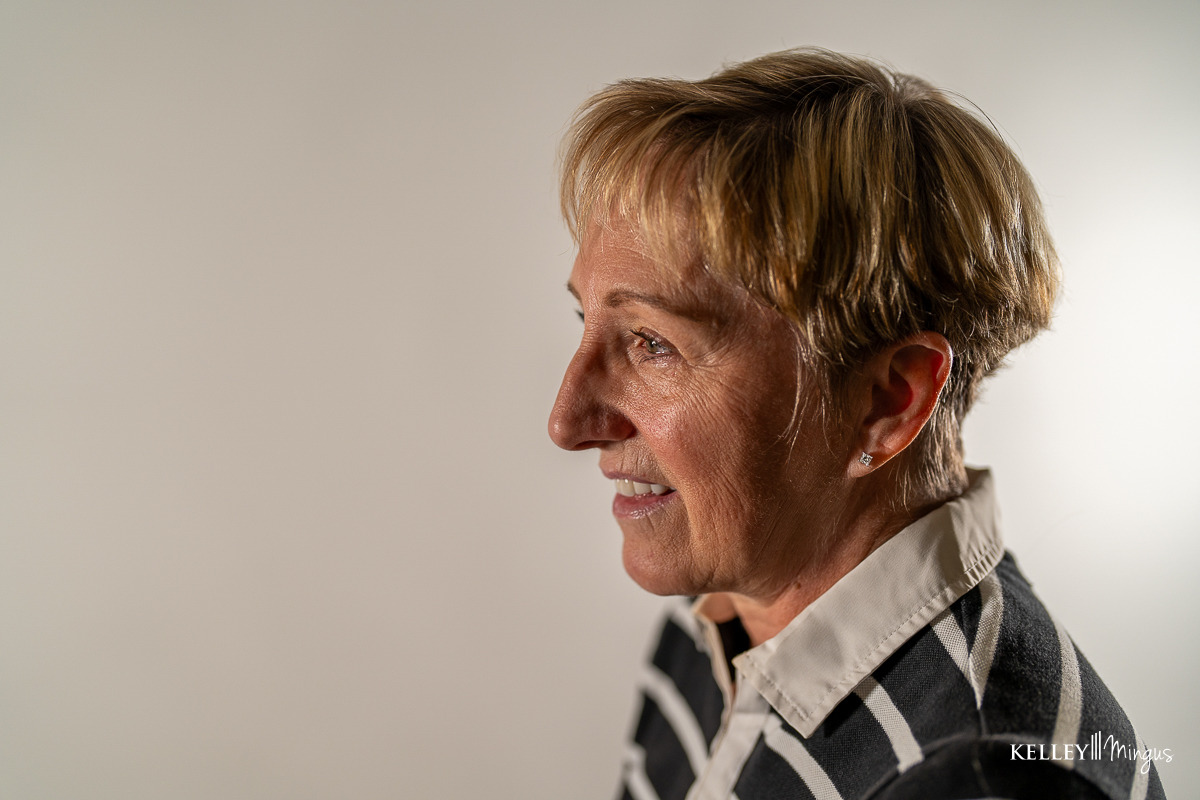 An older woman with short blond hair smiles, looking to the side, her bright smile reflecting confidence after teeth cleaning and cosmetic dentistry. She wears a striped shirt with a white collar. The background is plain and light-colored.