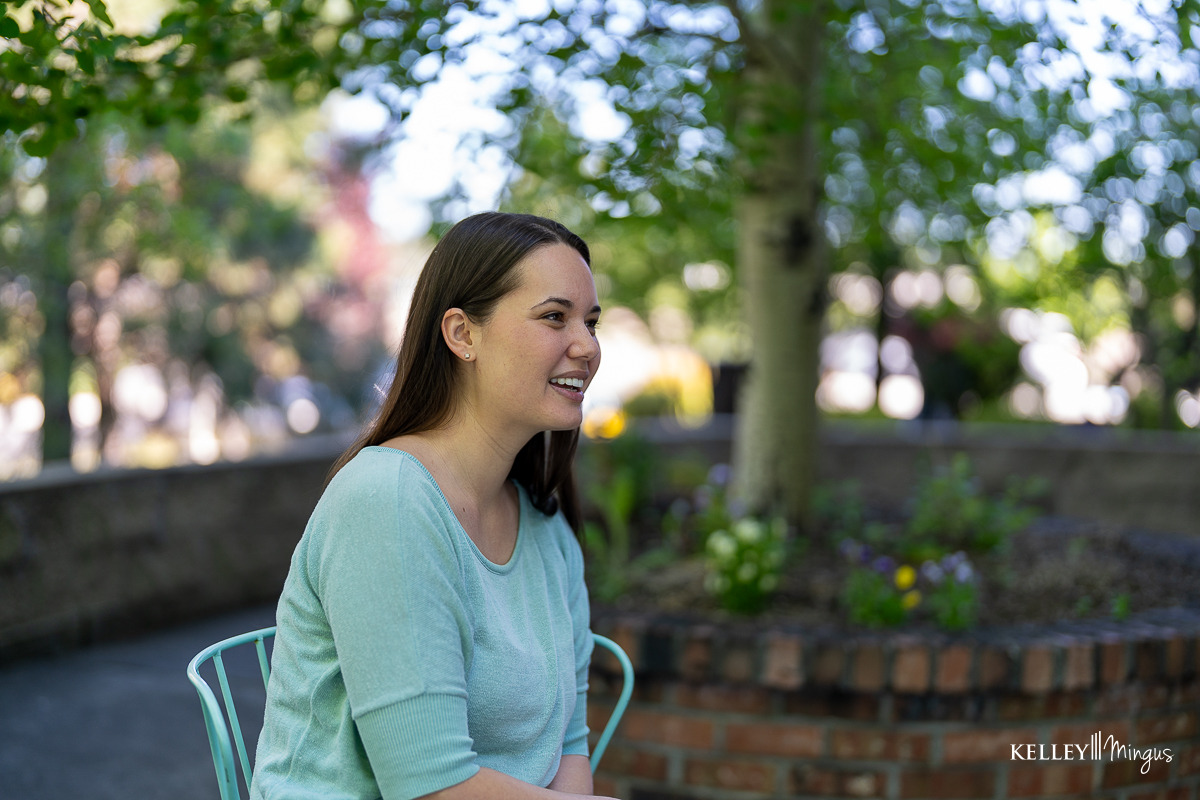A woman with long brown hair sits outdoors on a blue chair, smiling, her bright smile reflecting the confidence that comes from affordable cosmetic dentistry in Bend. She is wearing a light blue top, with trees and a brick planter in the background.