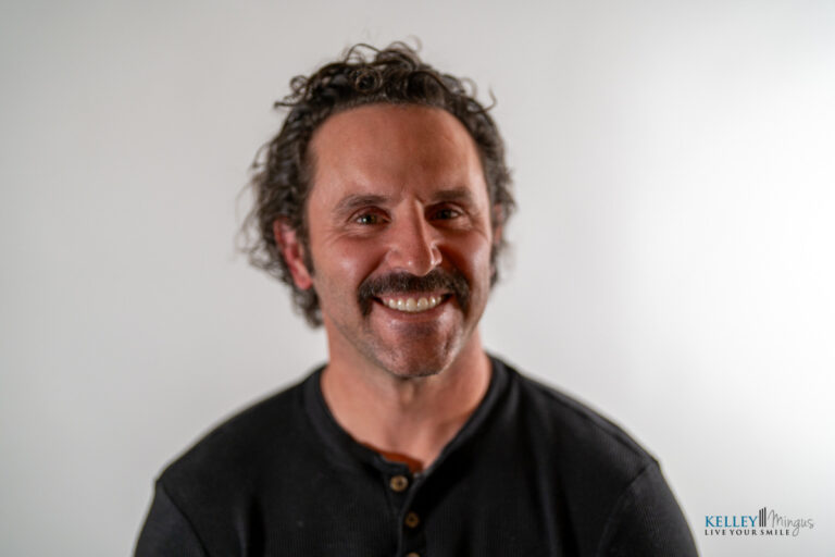 Man with curly hair and a mustache smiling, wearing a dark henley shirt, in front of a plain light background—resembling the approachable professionalism often associated with Kelley Mingus TMJ specialist.