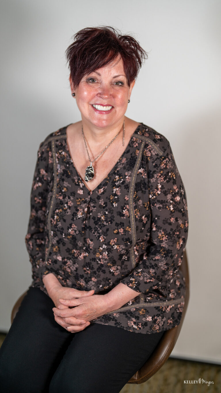 A woman with short, dark hair sits on a stool, smiling after teeth cleaning. She wears a dark floral top, black pants, necklace, and earrings against a plain light background.