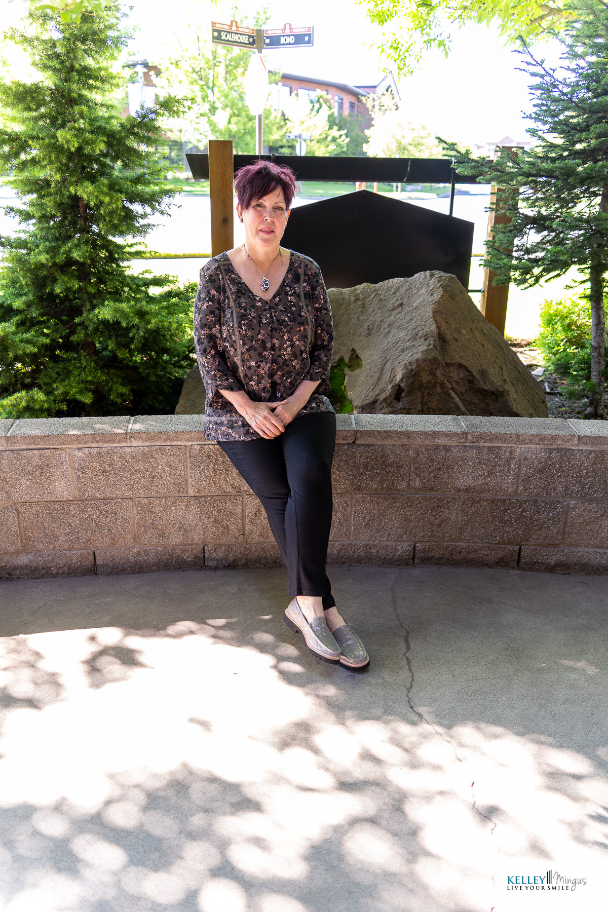 Woman with short hair sits on a stone bench outdoors, wearing a floral blouse, black pants, and slip-on shoes. With confidence that comes from her radiant smile—thanks to affordable cosmetic dentistry in Bend—she enjoys the fresh air among the trees and a large rock in the background.
