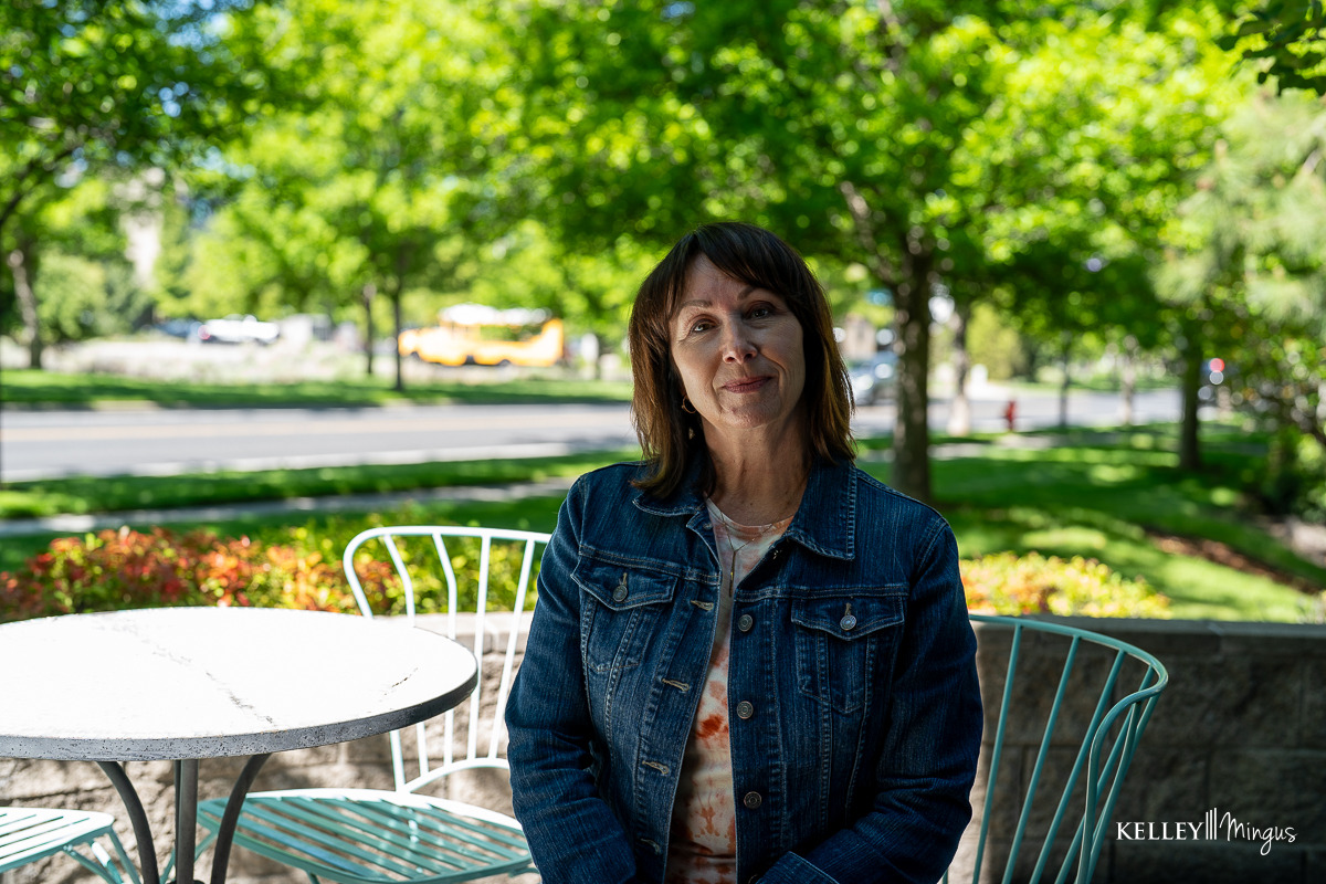 A woman in a denim jacket sits at a metal outdoor table in a shaded area with green trees and a road in the background, perhaps enjoying the peace after her visit to affordable cosmetic dentistry in Bend.
