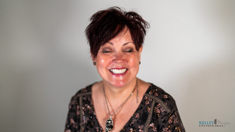 Woman with short dark hair and floral blouse smiles with eyes closed against a light background, radiating confidence often seen after Full Mouth Rehabilitation in Bend Oregon. She wears a pendant necklace, and there is a watermark in the bottom right corner.